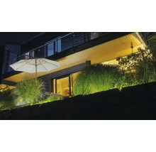 Beleuchtete moderne Terrasse bei Nacht mit weißem Sonnenschirm, Ziergräsern und warmer Außenbeleuchtung an einer Steinmauer.