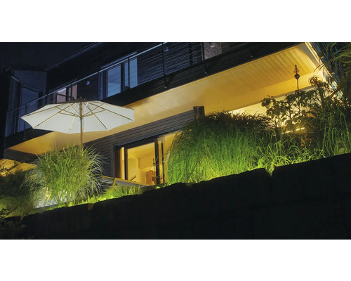 Beleuchtete moderne Terrasse bei Nacht mit weißem Sonnenschirm, Ziergräsern und warmer Außenbeleuchtung an einer Steinmauer.