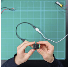 Eine Person hält ein kleines schwarzes Analog-Add-on-Elektronikmodul mit grünen Anschlüssen über einer Arbeitsfläche.