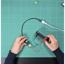 Hände montieren elektronische Bauteile und Kabelverbindungen mit Krokodilklemmen auf einer Arbeitsunterlage.