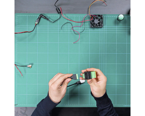 Hände verbinden ein Kabel mit einem SSR ADDON Modul auf einer Arbeitsfläche mit elektronischen Bauteilen wie Lüfter und Kabeln.