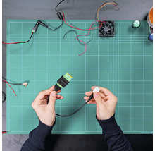 Elektronisches Anschlusskabel mit USB-Stecker und Klemmblock, gehalten in den Händen über einer Arbeitsmatte mit Elektronikkomponenten.
