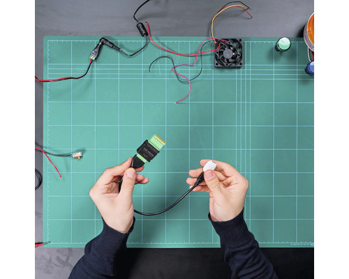 Elektronisches Anschlusskabel mit USB-Stecker und Klemmblock, gehalten in den Händen über einer Arbeitsmatte mit Elektronikkomponenten.