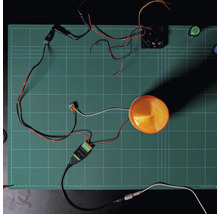 Shelly Solid State Relay Addon Modul mit angeschlossener Warnleuchte und Ventilator auf einer Schneidematte zur Demonstration der Verkabelung.