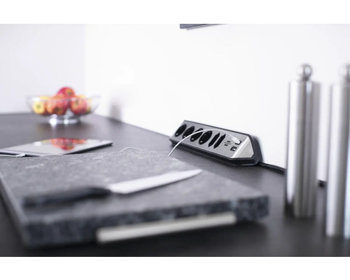 Szene mit einer Steckdosenleiste, einem Tablet, einem Schneidebrett, einem Messer und einer Schale mit Äpfeln auf einer Arbeitsplatte.