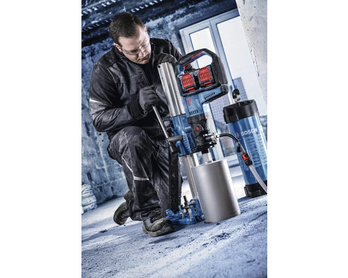 Handwerker bedient ein Bosch Akku-Diamantkernbohrgerät mit Bohrständer auf einem Betonboden in einer Baustellenumgebung.