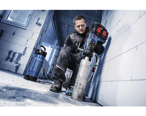 Handwerker nutzt ein Bosch Akku-Kernbohrgerät mit großem Bohraufsatz an einer Steinwand, verbunden mit einem Bosch Wasserbehälter auf einer Baustelle.