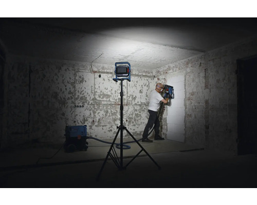 Ein Mann arbeitet mit einer Mauernutfräse in einem Raum, der von einem Baustrahler beleuchtet wird. Ein Staubsauger steht bereit.