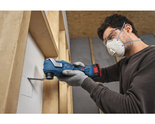 Person mit Schutzausrüstung schneidet mit einem Bosch Akkumulator-Multitool eine Öffnung in eine Wandbauplatte zwischen Holzständerwerk.