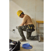 Ein Elektriker kniet und arbeitet an elektrischen Leitungen mit einem Werkzeugkoppel und einem gelben Schutzhelm.