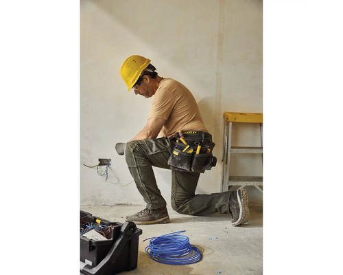 Ein Elektriker kniet und arbeitet an elektrischen Leitungen mit einem Werkzeugkoppel und einem gelben Schutzhelm.