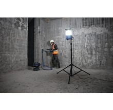 Ein Arbeiter bohrt mit einer Bohrmaschine ein Loch in eine Betonwand, unterstützt von einer Baustellenbeleuchtung und einem Staubsauger.