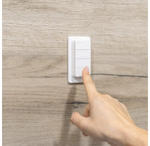 Weißer Funkwandschalter mit vier Tasten an einer Wand in Holzoptik, eine Hand bedient einen der Knöpfe.