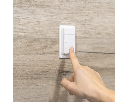 Weißer Funkwandschalter mit vier Tasten an einer Wand in Holzoptik, eine Hand bedient einen der Knöpfe.