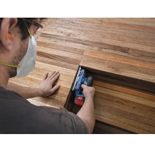 Handwerker bearbeitet eine Holzdiele mit einem Bosch Professional Akku-Hobel, trägt Schutzausrüstung wie Atemschutzmaske und Schutzbrille.
