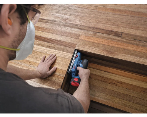 Handwerker bearbeitet eine Holzdiele mit einem Bosch Professional Akku-Hobel, trägt Schutzausrüstung wie Atemschutzmaske und Schutzbrille.