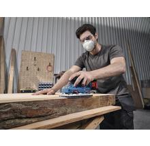 Ein Mann mit Atemschutzmaske und Schutzbrille bearbeitet eine Holzplatte mit einem Bosch Schwingschleifer in einer Werkstatt.