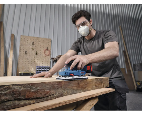 Ein Mann mit Atemschutzmaske und Schutzbrille bearbeitet eine Holzplatte mit einem Bosch Schwingschleifer in einer Werkstatt.