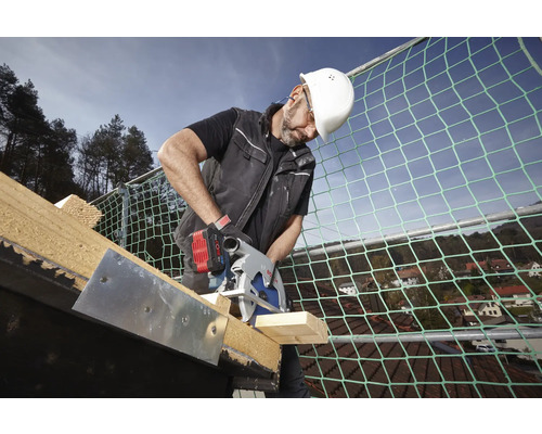 Handwerker mit weißem Helm schneidet mit einer Bosch Akku-Kreissäge Holz auf einem Dach mit Sicherheitsnetz.