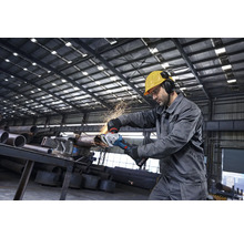 Ein Handwerker in Schutzkleidung bearbeitet ein Metallrohr mit einem Akku-Winkelschleifer in einer Industriehalle bei Funkenflug.