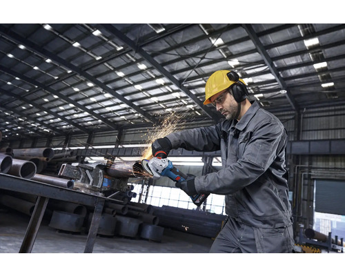 Ein Handwerker in Schutzkleidung bearbeitet ein Metallrohr mit einem Akku-Winkelschleifer in einer Industriehalle bei Funkenflug.