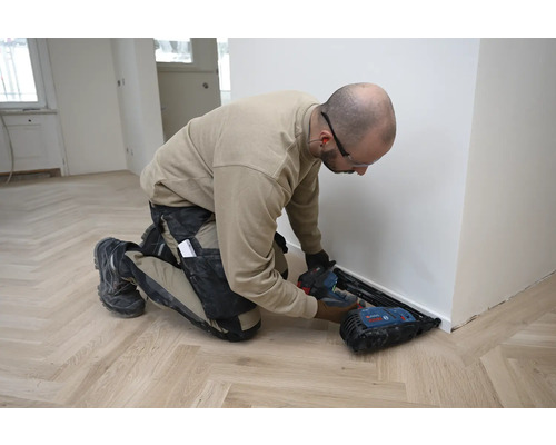 Handwerker montiert eine weiße Fußleiste mit einem batteriebetriebenen Nagler von Bosch auf einem hellen Parkettboden mit Fischgrätmuster.