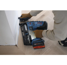 Eine Person befestigt mit einem Bosch Professional Akku-Nagler eine Sockelleiste an einer Wand über einem Holzfußboden im Fischgrätmuster.
