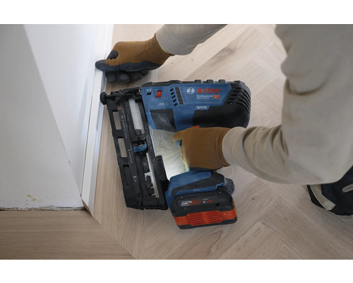Eine Person befestigt mit einem Bosch Professional Akku-Nagler eine Sockelleiste an einer Wand über einem Holzfußboden im Fischgrätmuster.