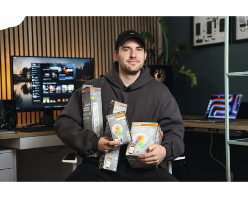 Mann im grauen Hoodie präsentiert Osram Smart Plus Beleuchtungsprodukte in einem modernen Büro mit Holzpaneelwand.