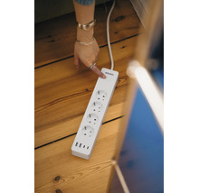 Osram smarte Steckdosenleiste mit vier Steckdosen und drei USB-Anschlüssen auf einem Holzboden. Eine Hand betätigt den Einschaltknopf.