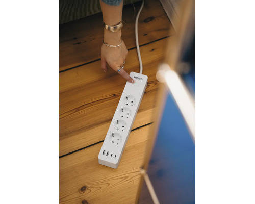 Osram smarte Steckdosenleiste mit vier Steckdosen und drei USB-Anschlüssen auf einem Holzboden. Eine Hand betätigt den Einschaltknopf.