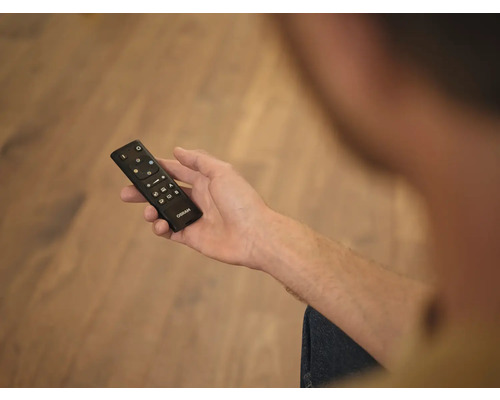 Eine Hand hält eine schwarze Fernbedienung der Marke Osram zur Lichtsteuerung über einem Fußboden aus Holz.