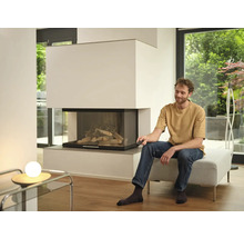 Ein Mann sitzt auf einem Hocker in einem hellen Wohnzimmer und bedient einen modernen Kamin mit Glasfront per Fernbedienung.