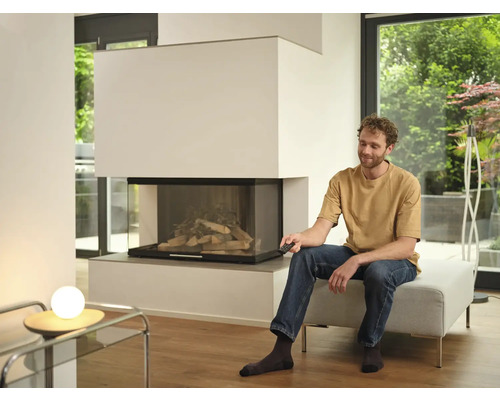 Ein Mann sitzt auf einem Hocker in einem hellen Wohnzimmer und bedient einen modernen Kamin mit Glasfront per Fernbedienung.