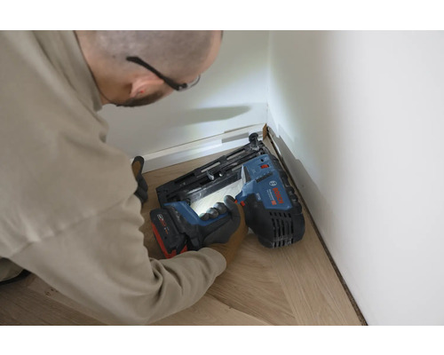 Person befestigt eine weiße Sockelleiste mit einem Bosch Akku-Nagler auf einem hellen Holzfußboden.
