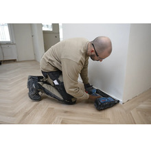 Handwerker montiert eine weiße Sockelleiste auf einem Holzboden mit Fischgrätmuster mit einem Akku-Nagler.