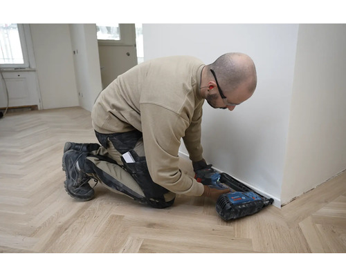 Handwerker montiert eine weiße Sockelleiste auf einem Holzboden mit Fischgrätmuster mit einem Akku-Nagler.