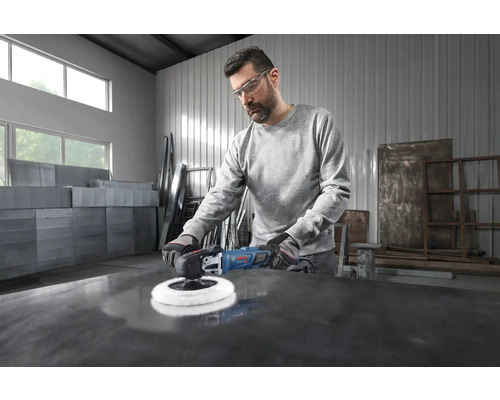 Ein Handwerker mit Schutzbrille poliert eine dunkle Metallfläche in einer hellen, geräumigen Werkstatt mit einer Bosch Poliermaschine.
