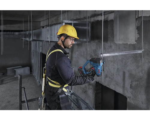 Ein Bauarbeiter mit Sicherheitsgurt und Helm benutzt ein Bosch Akku-Werkzeug an einer Metallstruktur auf einer Baustelle.