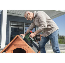 Ein Mann befestigt mit einem Bosch Akku-Tacker Dachpappe auf dem Dach einer hölzernen Hundehütte auf einer hellen Terrasse.