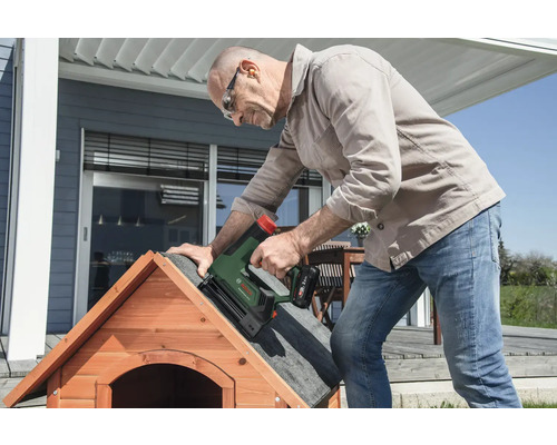 Ein Mann befestigt mit einem Bosch Akku-Tacker Dachpappe auf dem Dach einer hölzernen Hundehütte auf einer hellen Terrasse.