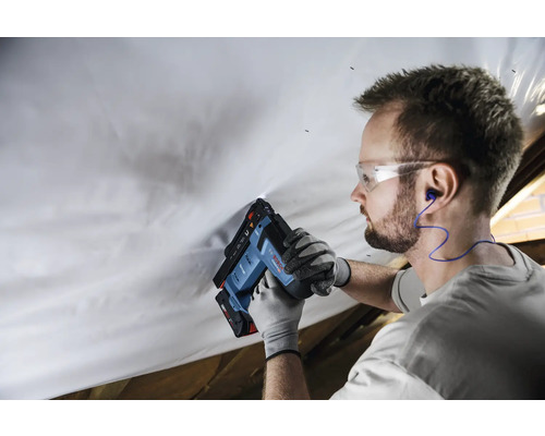 Handwerker befestigt Folie mit Bosch Akku-Tacker im Dachausbau, trägt Schutzbrille, Handschuhe und Gehörschutz.