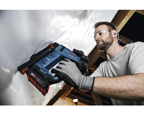 Handwerker mit Schutzausrüstung befestigt weiße Folie mit einem Bosch Professional Akku-Tacker an einer Dachkonstruktion aus Holz.