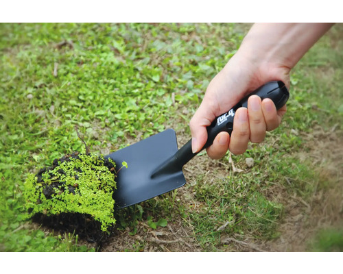 Eine Person hält eine schwarze Gartenschaufel mit einem Stück Erde und Pflanzen in der Hand.