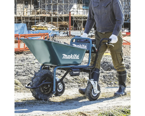 Eine Person schiebt eine mit Erde gefüllte Makita Akku-Schubkarre auf einer hellen Baustelle. Blaues Gerät mit drei Rädern und Makita Logo.