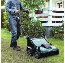 Eine Person mit Handschuhen und Stiefeln schiebt einen Makita Vertikutierer über einen Rasen in einem hellen, gepflegten Garten.