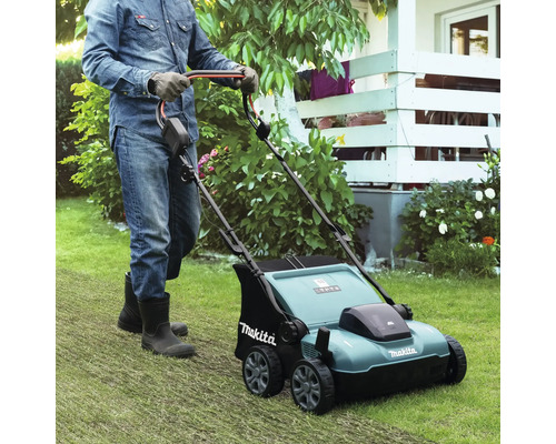 Eine Person mit Handschuhen und Stiefeln schiebt einen Makita Vertikutierer über einen Rasen in einem hellen, gepflegten Garten.