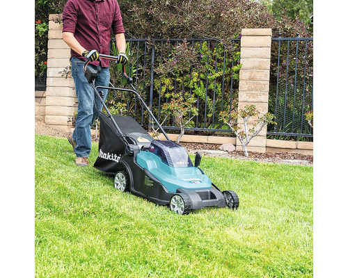 Eine Person verwendet einen batteriebetriebenen Rasenmäher von Makita auf einer gepflegten Rasenfläche vor einem Gartenzaun mit Steinmauer.