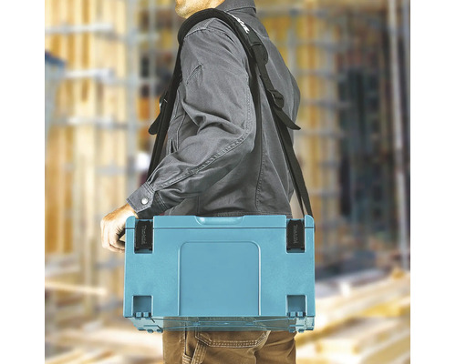 Eine Person trägt einen Makita Werkzeugkoffer an einem gepolsterten Schultergurt auf einer hellen Baustelle. Makita Logo.