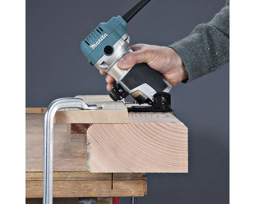 Eine Person bearbeitet die Kante eines auf einer Werkbank fixierten Holzbretts mit einer Makita Einhandfräse.